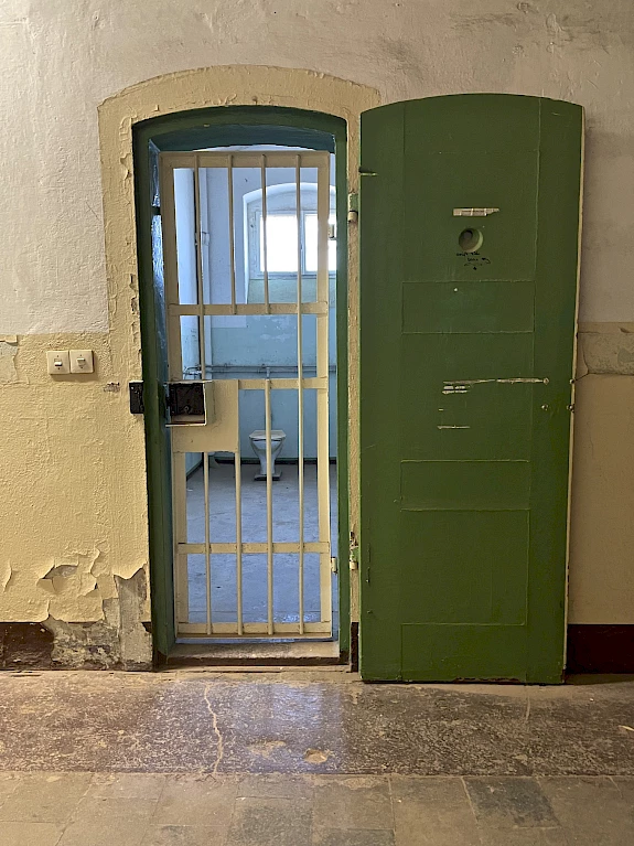 Blick auf eine geöffnete grüne Zellentür. Die sich dahinter befindenden Gitterstäbe, geben den Blick auf eine kleine Zelle mit Toilette und einem Fenster frei